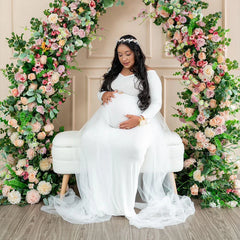 A pregnant woman wears a white long-sleeved maternity dress with a sheer mesh train.