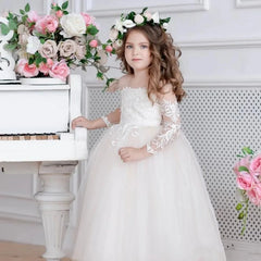 A young girl wears a white floor-length tulle ball gown with long lace sleeves and floral bodice.