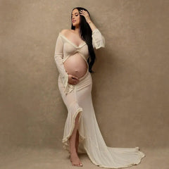 A pregnant woman wears a long-sleeved, off-the-shoulder sheer maternity gown with a front slit.