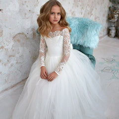 A young girl wears an ivory floor-length ball gown with long lace sleeves and a tulle skirt.