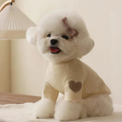 A small white dog wearing a beige long-sleeved shirt with a brown heart patch on the sleeve.
