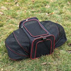 A black and red soft-sided pet carrier with expandable mesh sides sitting on grass.
