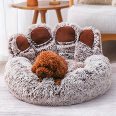A fluffy, bear paw-shaped pet bed in light brown with a small brown dog resting inside.
