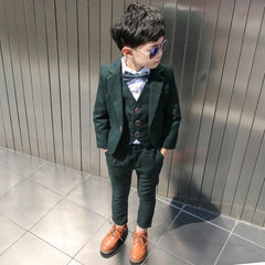A young boy wears a dark green four-piece formal suit with a blazer, vest, trousers, and bow tie.