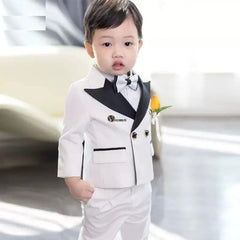 A young boy wears a white formal tuxedo suit with black lapels, a matching bowtie, and a yellow flower boutonniere.