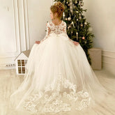 A girl wears a floor-length white lace flower girl dress with long sleeves and a bow at the back.