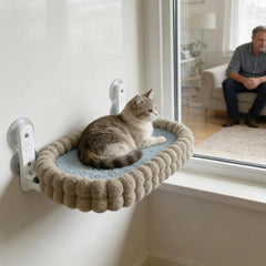 A cat rests on a plush, oval-shaped window perch attached to a glass pane with suction cups.