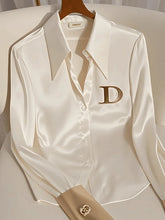 A white satin long-sleeve blouse with a turn-down collar and a gold letter D on the chest.