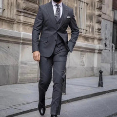 A man wears a grey three-piece slim-fit suit with a peaked lapel and a patterned tie.