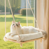 A fluffy white cat rests on a wooden window perch hammock with a soft plush cushion.