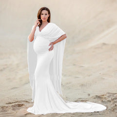 A pregnant woman wears a white V-neck mermaid maternity gown with batwing sleeves on a sandy beach.