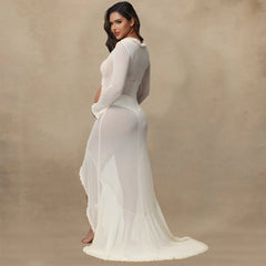 A pregnant woman wears a long-sleeved white sheer maternity gown with a ruffled hem and side slit.