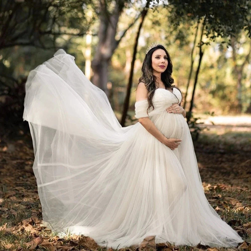 A pregnant woman wears an off-shoulder white tulle maternity gown in a forest setting.