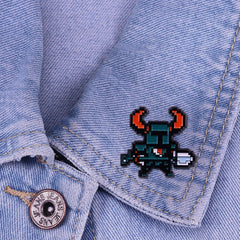 A pixel art enamel pin of a dark blue knight with red horns attached to a denim jacket.