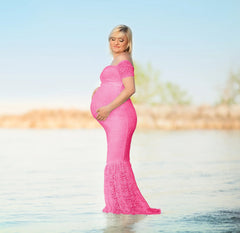 A pregnant woman stands in shallow water wearing a form-fitting pink lace maternity gown.