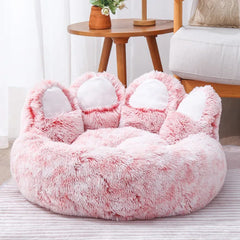 A fluffy, pink, bear paw-shaped pet bed with white toe pads sits on a striped floor.