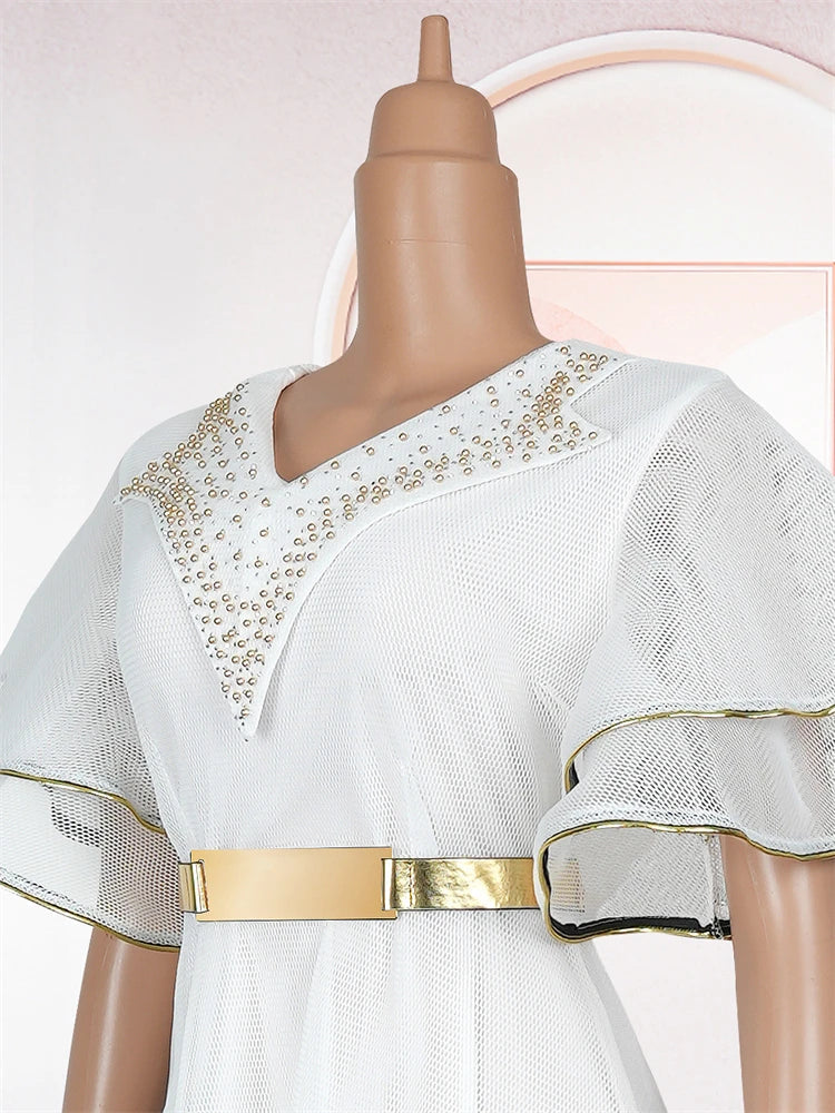 A white mesh maxi dress featuring gold-trimmed ruffle sleeves, a metallic belt, and beaded neckline.