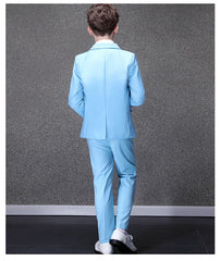 A boy stands facing away wearing a sky blue formal suit with a notched collar and white shoes.