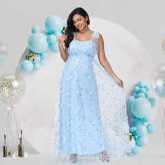 A pregnant woman wears a light blue sleeveless tulle gown with floral embroidery and strap details.