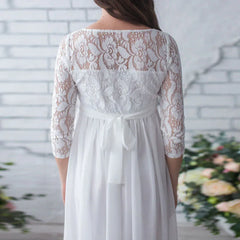 A white maternity maxi dress with a lace bodice, three-quarter sleeves, and a tie-back sash.