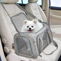 A small white dog sits inside a grey, soft-sided mesh pet carrier on a car seat.
