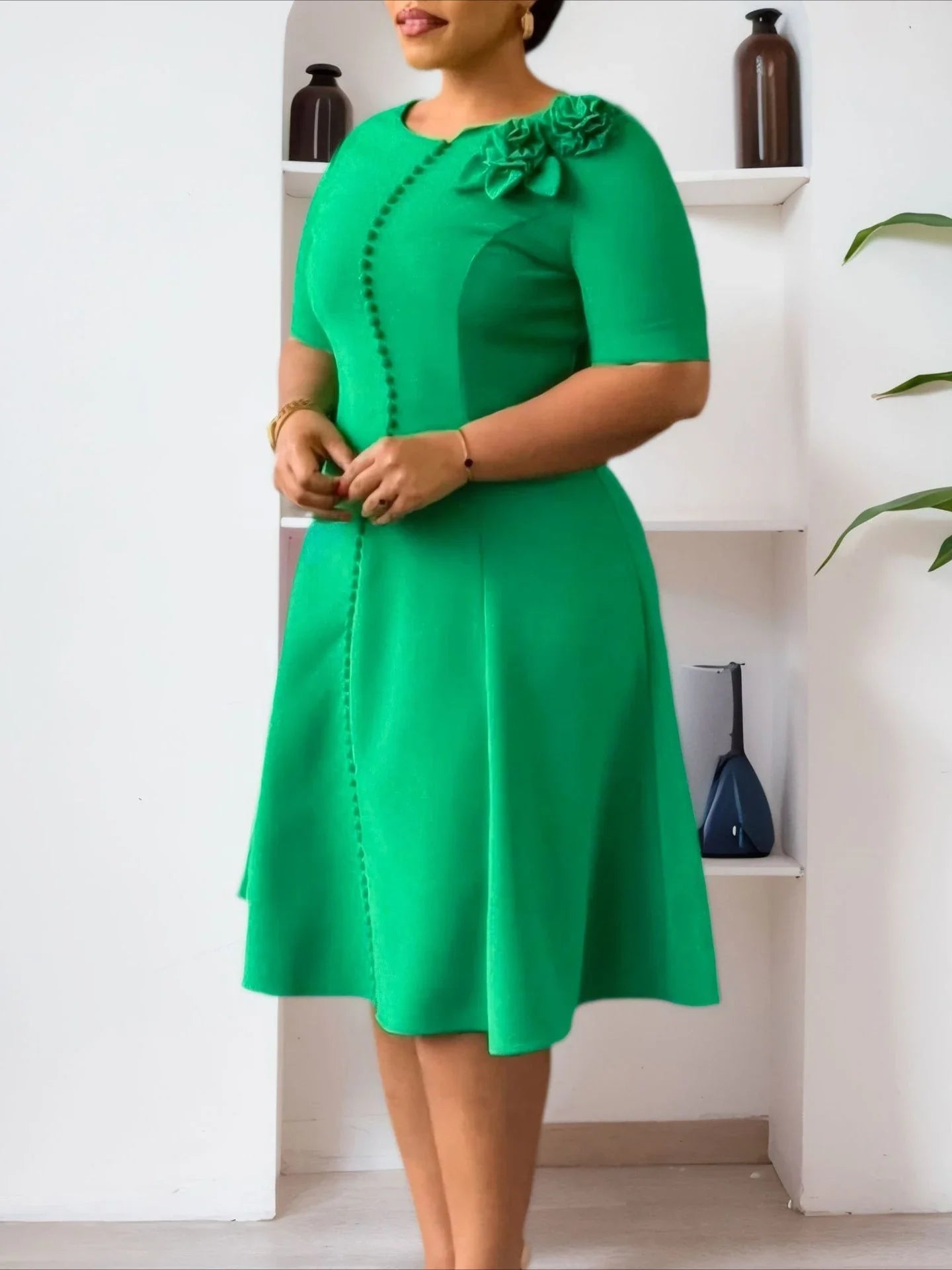 A woman wears a knee-length green A-line dress with short sleeves, button details, and floral trim.