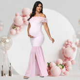 A pregnant woman wears a light pink off-shoulder maternity gown with a floral neckline and split hem.