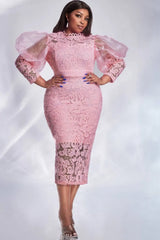 A woman wears a pink lace midi dress with sheer puff sleeves and a slim sheath silhouette.