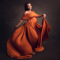 A pregnant woman wears a flowing, off-the-shoulder, ankle-length rust-colored satin maternity dress.