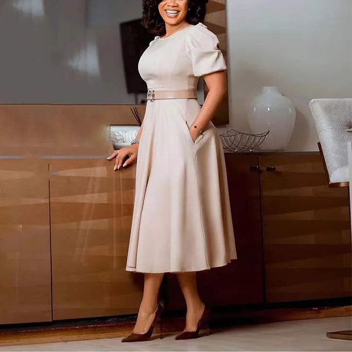 A woman wears a beige short-sleeved midi dress with a belted waist and pleated A-line skirt.