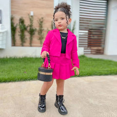 A young girl wears a bright pink long-sleeve jacket and matching pleated miniskirt set.