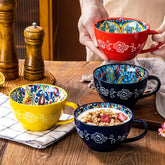 A person holds a red ceramic mug above a wooden table featuring yellow, dark blue, and patterned mugs.