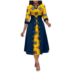 A woman wears a mid-calf A-line dress with a yellow Ankara print and a navy blue buttoned overlay.
