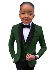 A young boy wears a dark green slim-fit suit jacket with black lapels, a vest, and a bowtie.