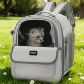 A light gray pet carrier backpack with a clear front window containing a cat, set on green grass.