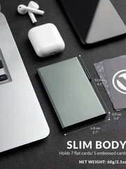 A slim, metallic gray RFID-blocking card holder sits on a desk next to a laptop and earbuds.