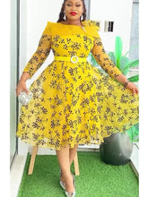 A woman wears a yellow knee-length dress with a black floral print, long sleeves, and a belt.