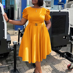 A woman wears a mustard yellow, short-sleeved, mid-calf pleated dress with a belted waist.