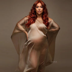 A pregnant woman wears a sheer, ankle-length, boat-neck maternity cape in a soft beige satin.