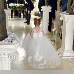 A young girl wears a white floor-length ball gown with lace detailing and a large back bow.