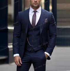 A man wears a navy blue three-piece suit with a white shirt, patterned tie, and pocket square.