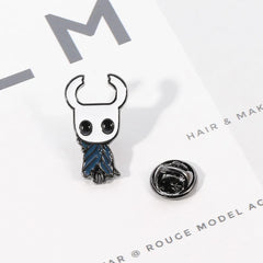 A Hollow Knight enamel pin featuring the character with a blue cloak and a metal backing clutch.