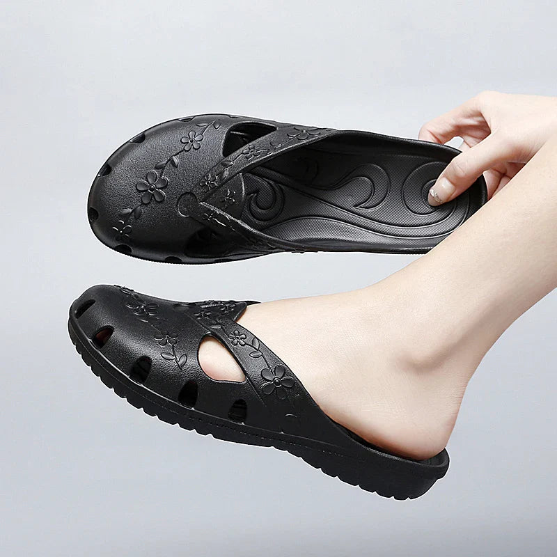 A hand holds a black, floral-patterned jelly slipper with cutouts and a flat sole.