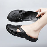 A hand holds a black, floral-patterned jelly slipper with cutouts and a flat sole.