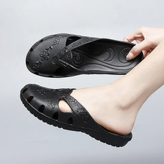 A hand holds a black, floral-patterned jelly slipper with cutouts and a flat sole.