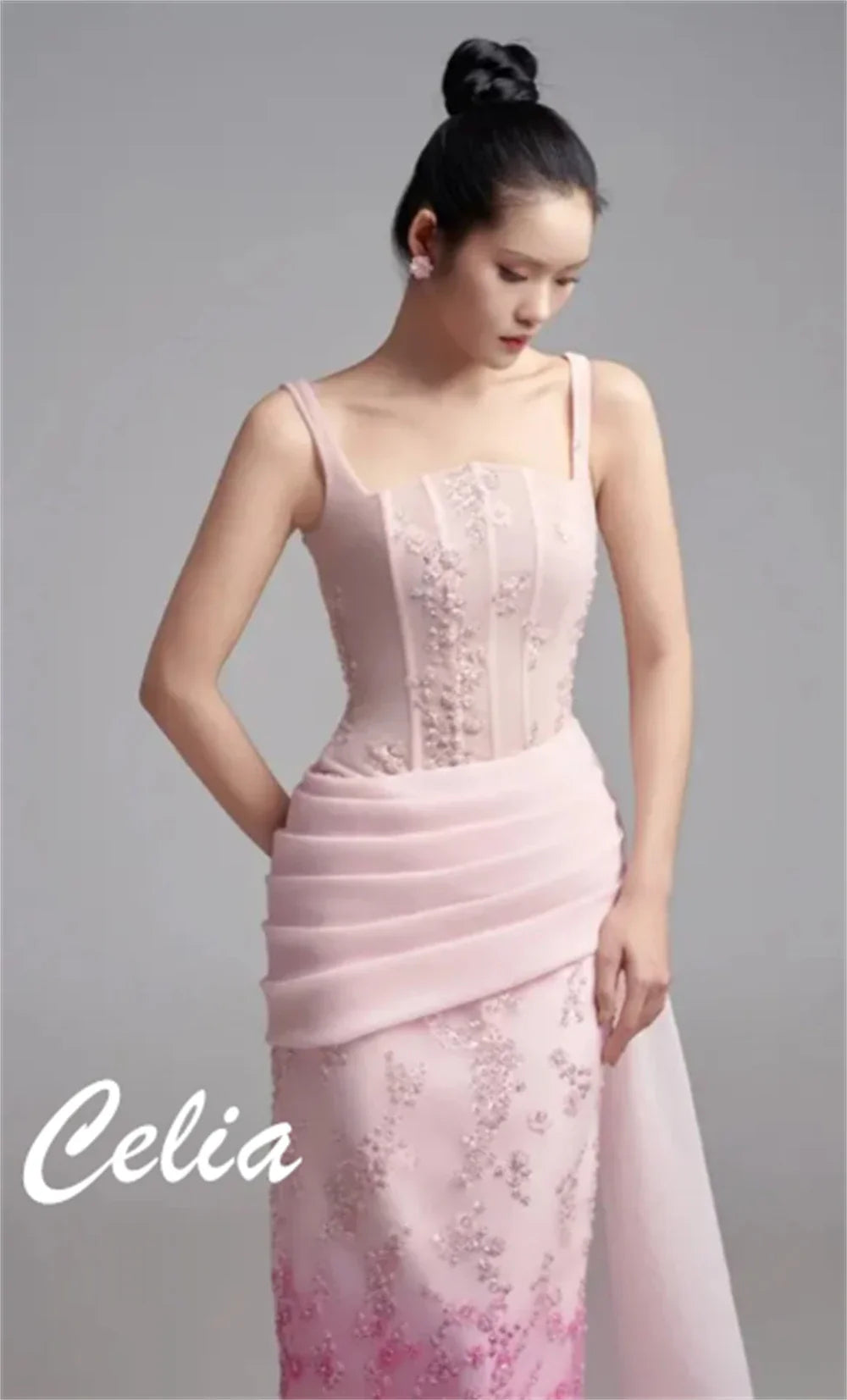 A model wears a sleeveless pink floor-length evening gown with floral embroidery and pleated waist.