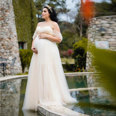A pregnant woman wears an off-shoulder white chiffon maxi dress for a photoshoot.
