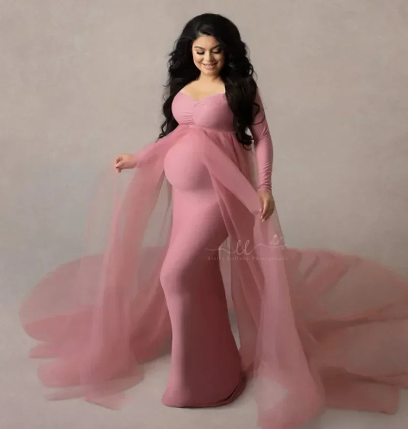 A pregnant woman wears a long-sleeved, dusty pink maternity dress with a sheer tulle cape train.