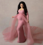 A pregnant woman wears a long-sleeved, dusty pink maternity dress with a sheer tulle cape train.