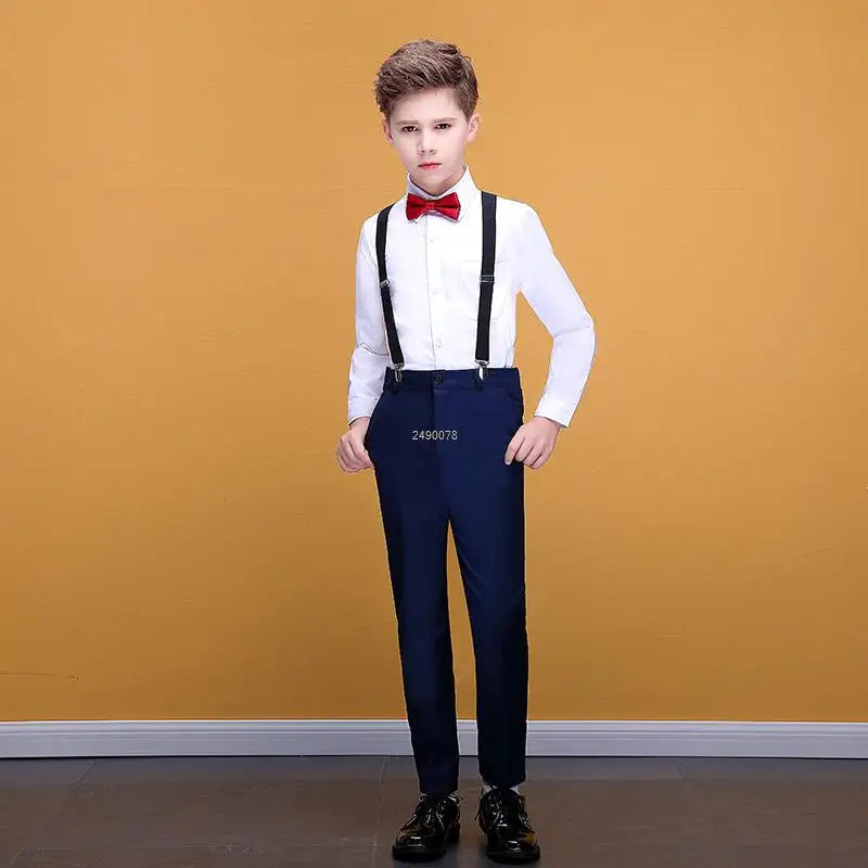 A boy wears a formal outfit consisting of a white button-down shirt, a red bow tie, black suspenders, and navy blue trousers.